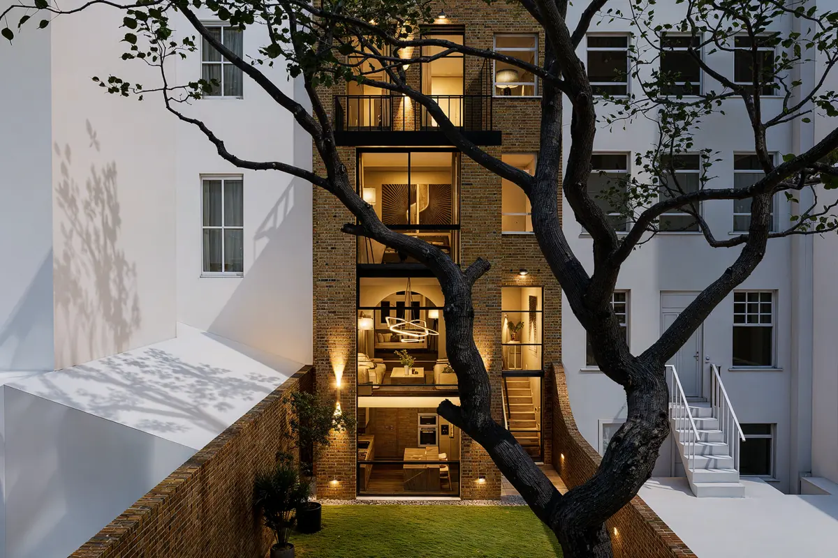 CGI render at dusk of the proposed rear elevation: full-height glazed four-storey rear with lit interiors, garden and tree in the foreground
