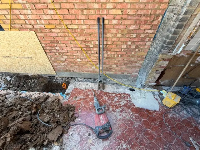 Foundation trial-pit excavation along the rear wall with breaker and post-hole tools, tiled floor of old extension