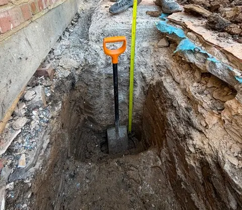 Deep trial pit beside the house wall with spade and tape measure showing foundation depth