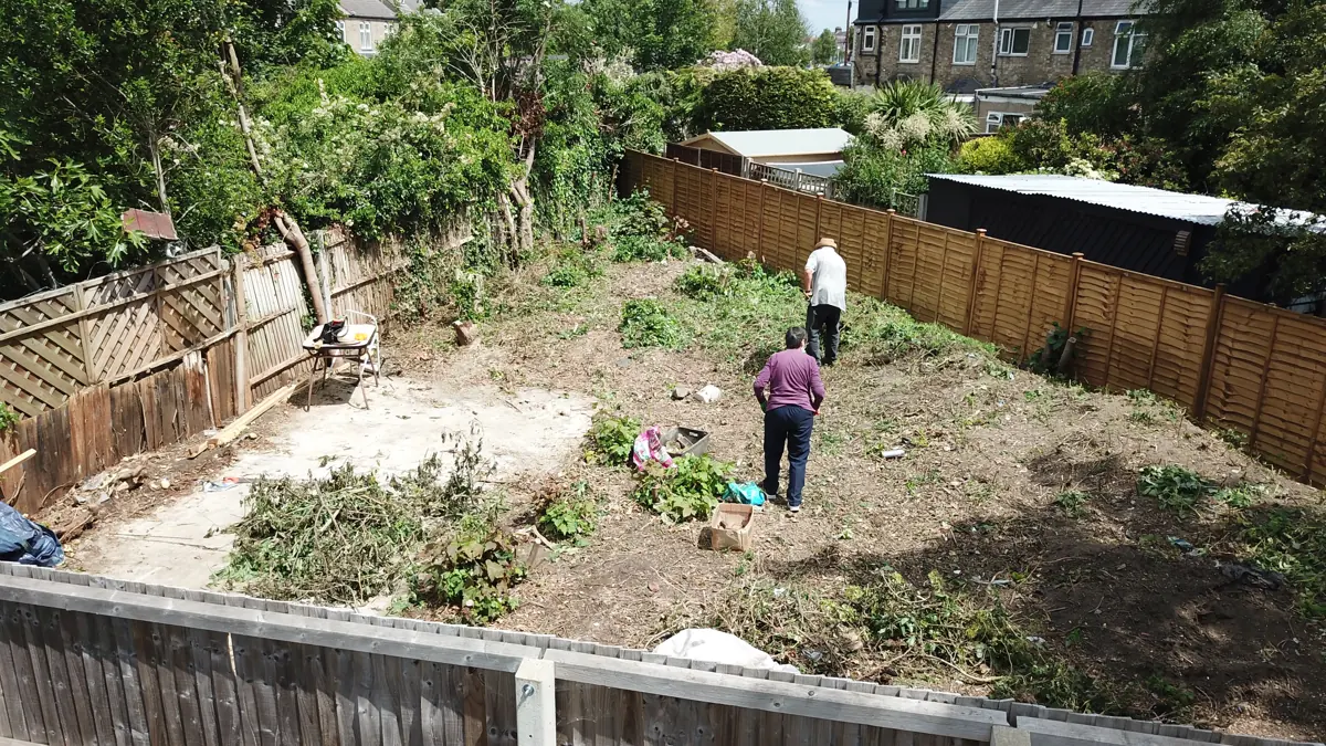 Low drone view across the cleared plot during site clearance, with timber fences and the backs of neighbouring houses
